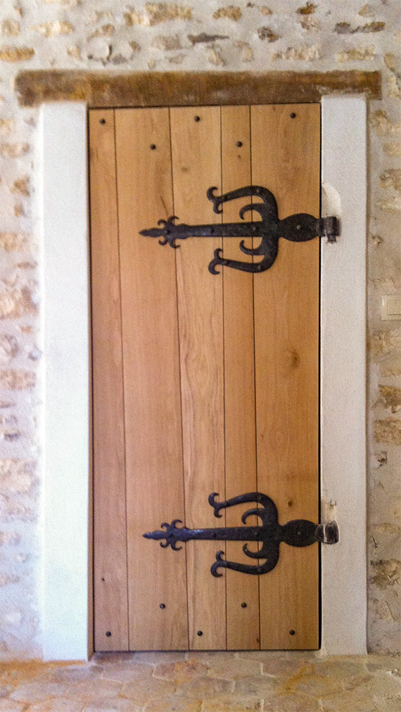Vue d'une porte créée sur mesure dans un style médiéval avec des planches de tailles irrégulières, des ferronneries stylisées et de gros clous en fer forgé